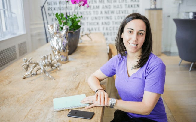 Brunette girl in purple shirt sitting at table smiling.