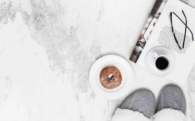 Grey slippers styled beside a bagel on a plate, with a cup of coffee on top of a magazine