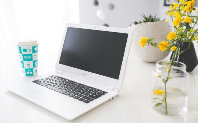 A laptop on a desk with yellow flowers in a vase, and a cup of coffee beside it