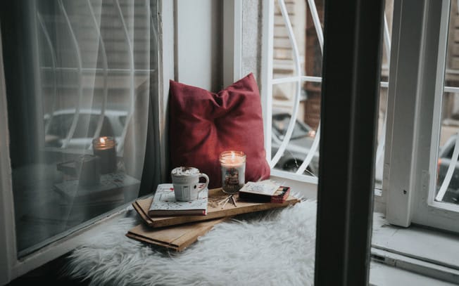 Pillow, candle and hot chocolate on top of a fuzzy blanket looking out a window