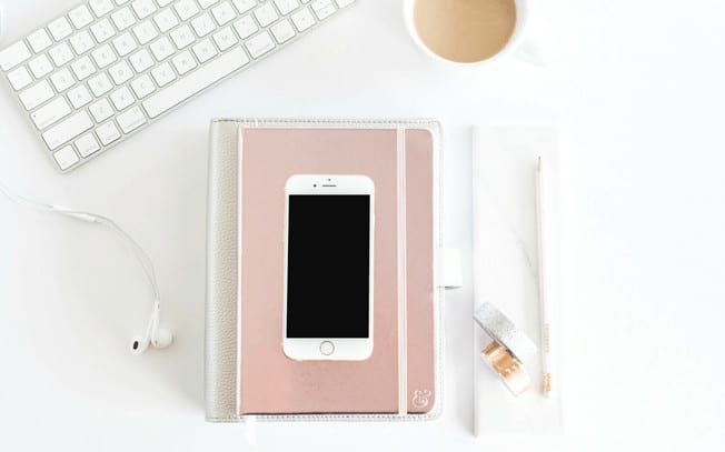 An iPhone on top of a notebook beside a cup of coffee and a keyboard