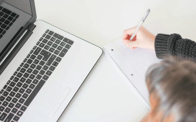 girl writing on paper at her laptop