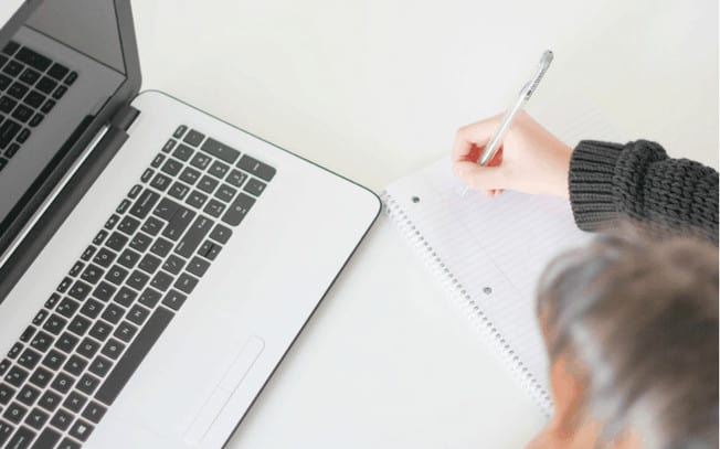 Person writing on a piece of paper at her laptop