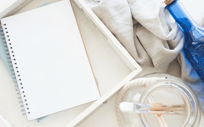 A styled photo of notepad, paintbrushes, blanket and blue bottl