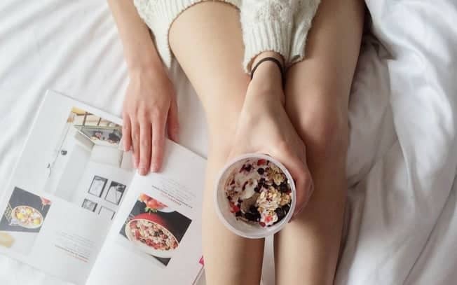 Person in bed reading a magazine and holding food