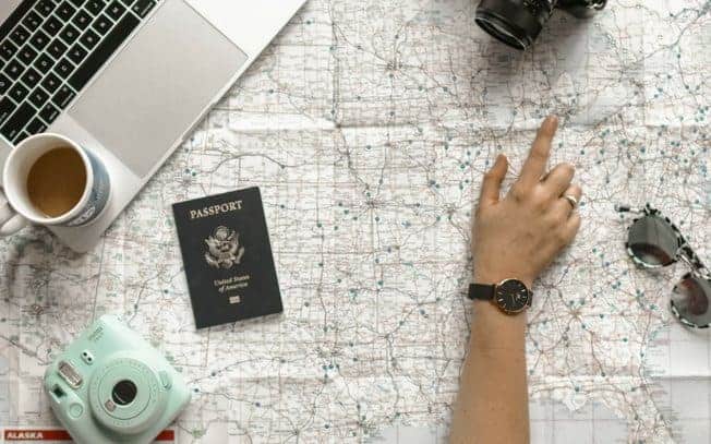 hand pointing to map beside passport, laptop, coffee, sunglasses and camera