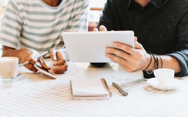 Two people using an iPad and a notebook. Coffee on a table.