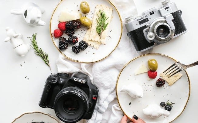 two cameras beside a plate of crackers, cheese, olives and fruit