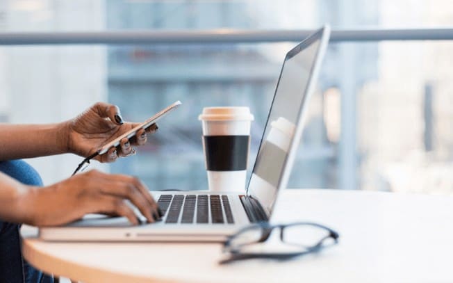 A person typing at a laptop and holding their phone