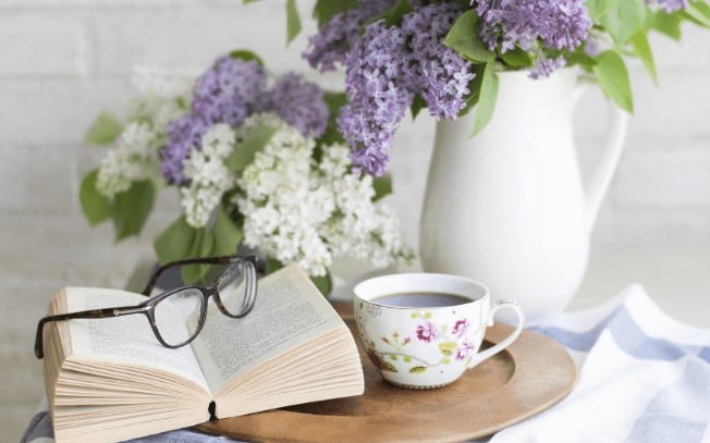 A book on a table with white and purple flowers, beside a cup of tea and glasses laying on top of the book.