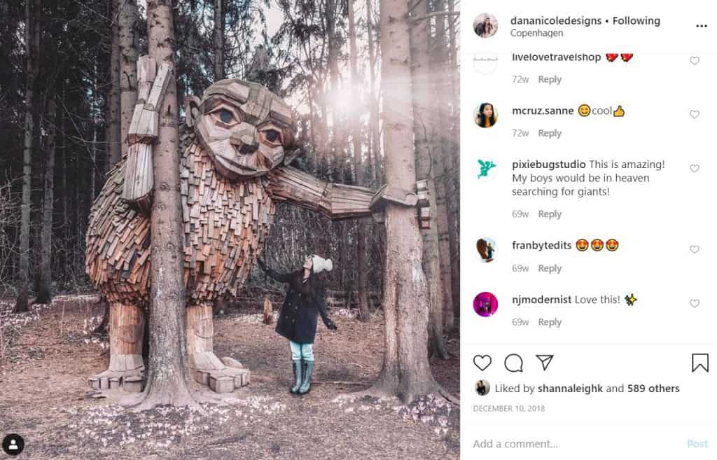 Screenshot of an Instagram feed image where a girl is looking up at a giant wooden statue of a giant in the woods