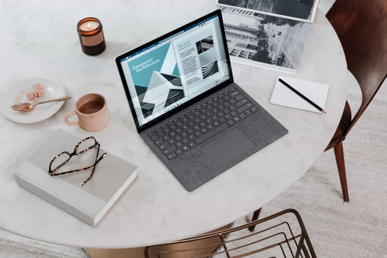 A laptop on a white table next to a candle, a pink cup of coffee, a book with glasses on top, and a paper with a pen.