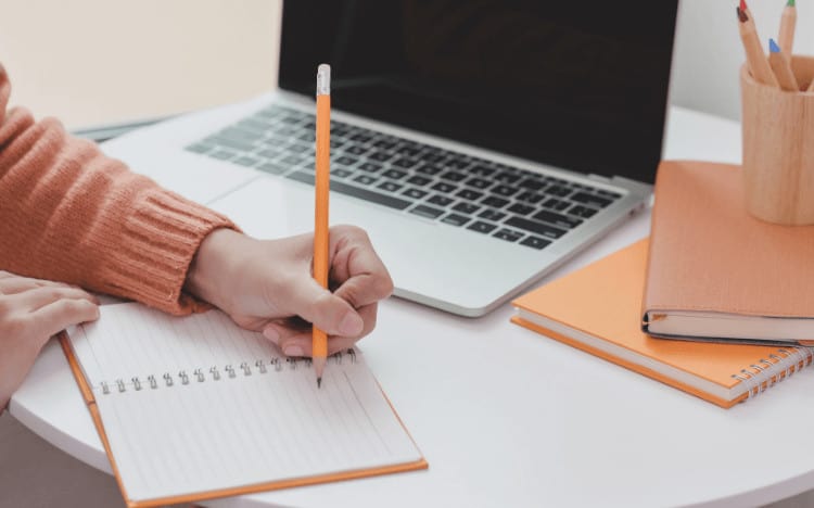 A woman writing in a notebook with a laptop beside her.